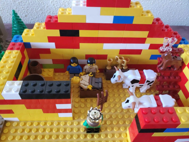 Auf dem Bild ist zu sehen, wie eine Minifigur in einer Krippe in einem Stall liegt, um sie herum stehen Maria und Josef, ein König sowie zwei Ochsen.